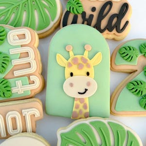 Könnte beinhalten: Auswahl an dekorierten Zuckerkeksen mit Dschungel-Thema. Die Kekse zeigen eine Giraffe, tropische Blätter und die Worte "two wild". Die Kekse sind mit grüner, gelber und weißer Glasur verziert.