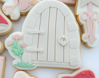 Op de afbeelding: Assortiment van versierde suikerkoekjes met een feeënthema. Koekjes omvatten een witte deur met bloemaccenten, een fee, een sterrenstaf, het nummer één en roze paddenstoelkoekjes.