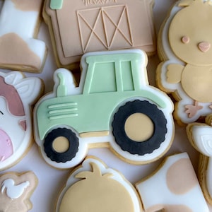 Könnte beinhalten: Eine Sammlung dekorierter Zuckerkekse zum Thema Bauernhof. Die Kekse umfassen einen hellgrünen Traktor, eine Scheune, ein Küken und eine Kuh. Die Kekse sind mit weißem, hellgrünem, beigem und schwarzem Zuckerguss verziert.