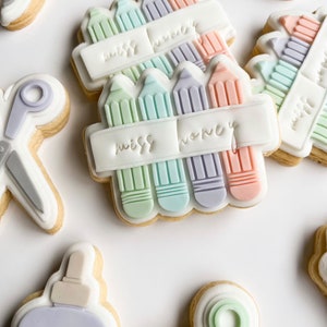 Puede incluir: Un conjunto de seis galletas decoradas. Las galletas están decoradas con glaseado de color pastel y presentan imágenes de tijeras, botellas de pegamento y lápices. Los lápices están dispuestos en una pila y tienen una pancarta que dice "miss nancy".