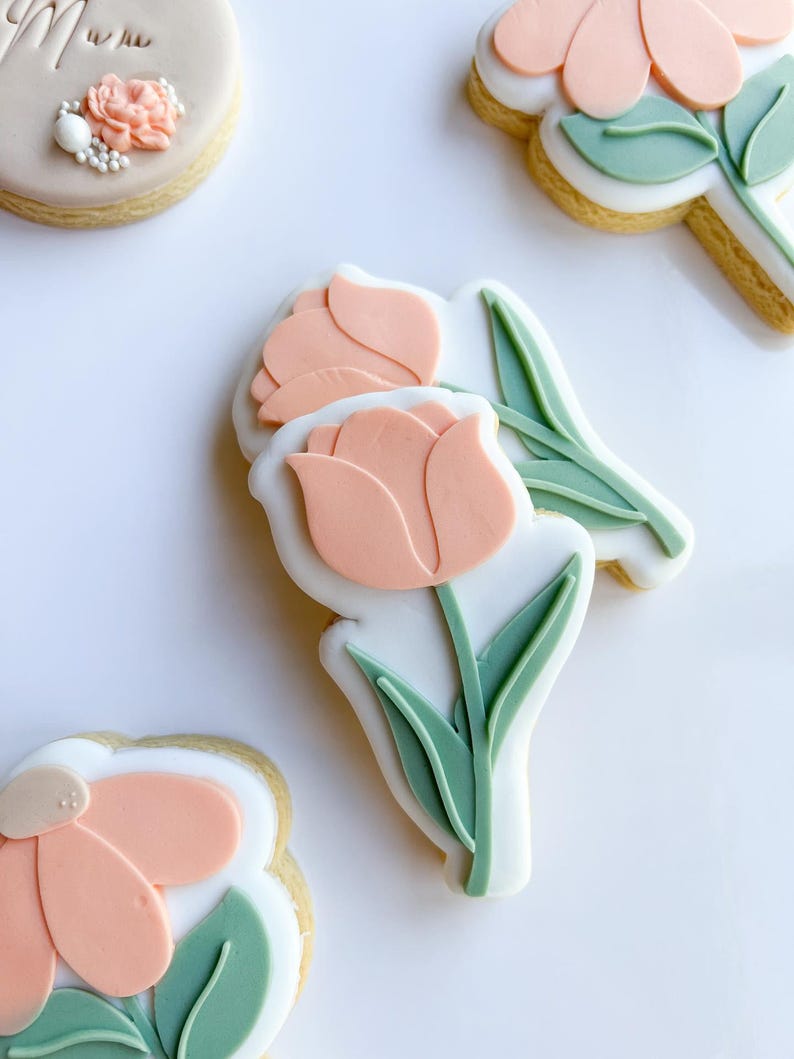 Puede incluir: Tres galletas de az&uacute;car decoradas con glaseado rosa pastel y verde. Las galletas tienen forma de flores con tallo y hojas.