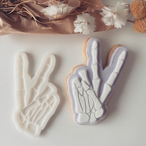Może przedstawiać: Dwa białe plastikowe wykrawacze do ciasteczek w kształcie znaku pokoju z wzorem szkieletowej dłoni. Wykrawacze do ciasteczek znajdują się na białej powierzchni.