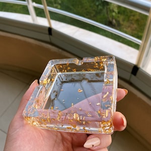 Peut inclure: Cendrier carré transparent avec des flocons de feuilles d'or incrustés dans la résine. Le cendrier a une forme rectangulaire avec des bords surélevés et de petites encoches pour poser les cigarettes. Le cendrier est tenu dans une main.
