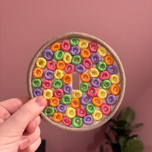 May include: A round, gray, ceramic coaster with a colorful pattern of cereal rings. The rings are in shades of yellow, orange, pink, purple, and green.