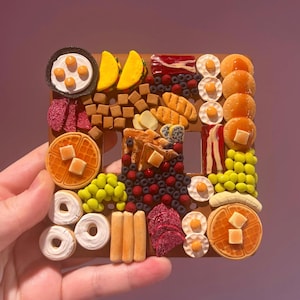 May include: A square light switch cover plate decorated with miniature food items including waffles with butter, pancakes with syrup, bacon strips, fried eggs, donuts, tacos, sausages, berries, and various small brown cubes on a brown base.