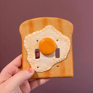 Peut inclure: Un cache-interrupteur fantaisie en forme de tranche de pain grillé avec un motif d'œuf au plat. Le pain grillé est de couleur brun clair avec des bords plus foncés, et l'œuf a un jaune d'œuf jaune et des blancs d'œufs blancs. Il comporte deux ouvertures pour les interrupteurs.