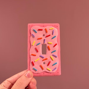 May include: A pink light switch cover decorated to resemble a frosted donut with colorful sprinkles. The cover has a rectangular shape with a cutout for the switch and is held against a pink background.