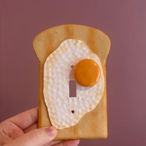 May include: A decorative light switch cover in the shape of a slice of toast with a fried egg on top. The toast is a light brown colour, and the egg has a white base with a yellow yolk. The switch opening is a vertical rectangle.