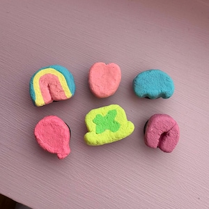 May include: Six colorful clay ornaments in various shapes, including a rainbow, a heart, a horseshoe, a hat, and two other abstract shapes. The ornaments are all pink, blue, yellow, and green.