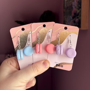 Könnte beinhalten: Drei Paar Macaron-Ohrringe auf Präsentationskarten. Die Macarons sind in Blautönen, Rosa und Lila. Die Karten haben ein gold-rosa Design und den Text "@lilclawkitchen".