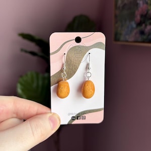 Può includere: Un paio di orecchini pendenti con piccoli ciondoli marroni a forma di patata. Gli orecchini sono esposti su una carta rosa e dorata con il testo "@lilcloudcraft".