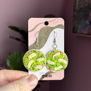 Puede incluir: Un par de pendientes de plata con un diseño redondo, verde y blanco con temática de comida. Los pendientes están en una tarjeta blanca con el texto "@lilcloudkitchen" y los iconos de las redes sociales.