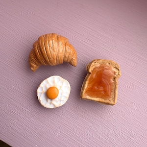 Könnte beinhalten: Drei Miniatur-Lebensmittel: ein Croissant, ein Stück Toast mit Marmelade und ein Spiegelei. Die Artikel sind aus Ton gefertigt und in realistischen Farben bemalt.