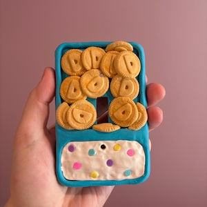 Op de afbeelding: Een grillige lichtschakelaar, versierd met een speels ontwerp. De afdekking is helder turquoise, versierd met miniatuur koekjesvormige versieringen. Het onderste gedeelte heeft een witte, frosting-achtige textuur met kleurrijke stippen.
