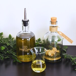 Può includere: Due bottiglie di vetro con liquido trasparente e un piccolo bicchiere pieno di un liquido giallo. La bottiglia a sinistra ha un'etichetta che recita "Rhum'fluence DIY Rum". La bottiglia a destra ha un'etichetta di carta marrone che recita "Polvere di spezie per pan di zenzero".