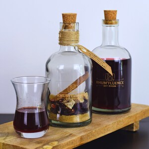 Può includere: Due bottiglie di vetro trasparente con tappi di sughero e un piccolo bicchiere pieno di un liquido rosso scuro. Le bottiglie sono etichettate "RHUM'FLUENCE DIY RHUM". Le bottiglie e il bicchiere sono su un vassoio di legno.