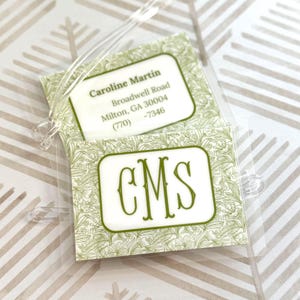 May include: A clear plastic luggage tag with a green and white floral pattern. The tag has two sections with text: "Caroline Martin" and address details, and "CMS" in a green frame.