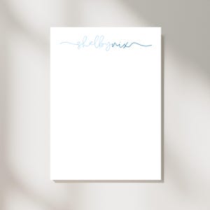 May include: A white notepad with the name "shelbynix" written in a delicate, light blue script across the top. The notepad is rectangular and sits on a neutral background.