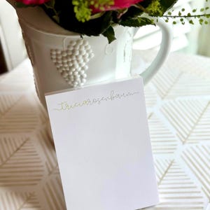 May include: A white notepad with the name "Tricia Rosenbaum" in gold script is in front of a white jug with a grape design. The jug holds pink roses and green foliage. The background has a geometric pattern.