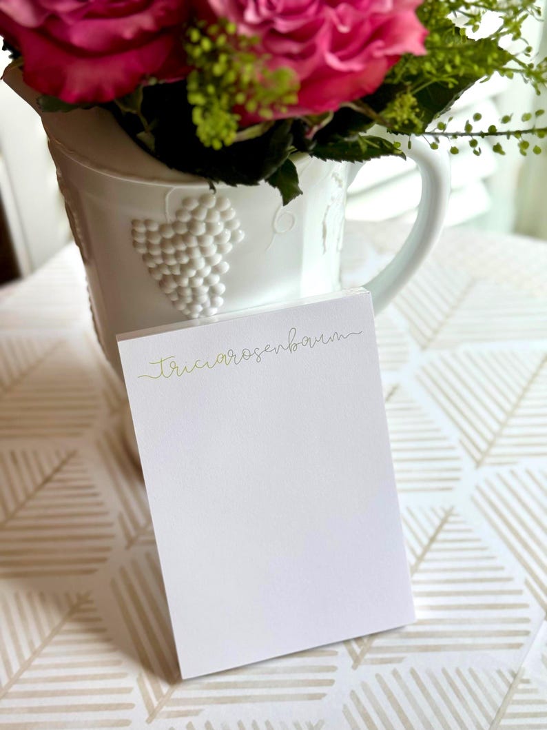 May include: A white notepad with the name "Tricia Rosenbaum" in gold script is in front of a white jug with a grape design. The jug holds pink roses and green foliage. The background has a geometric pattern.