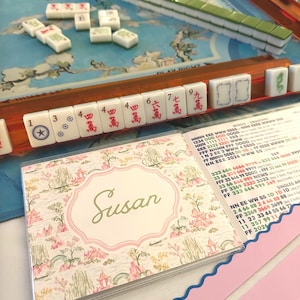 May include: A Mahjong set featuring a blue game board with floral patterns, green and white tiles, and an orange tile rack. A personalized card holder displays the name Susan in cursive script over a pink and white landscape illustration.