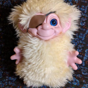 May include: A plush troll doll with long, shaggy cream-colored synthetic fur, a pink plastic face, and one bright blue eye. The doll features a brown faux leather eye patch covering the left eye and has small, outstretched pink plastic hands.