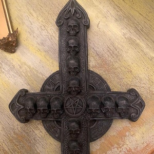 Op de afbeelding: Een zwarte metalen wanddecoratie in de vorm van een omgekeerd kruis met een pentagram in het midden. Het kruis is versierd met schedels en ingewikkelde details.