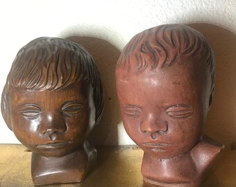 May include: Two ceramic sculptures of children's heads with closed eyes. The sculpture on the left is brown and the sculpture on the right is reddish brown.
