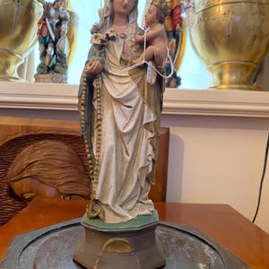 Puede incluir: Estatua religiosa de la Virgen María con el niño Jesús. La figura está pintada en blanco, dorado y verde, con un rosario y una corona. Se encuentra sobre una base de madera oscura, probablemente decorativa.