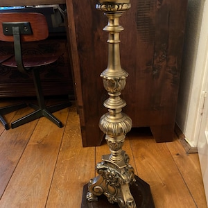 Può includere: Un alto e ornato candelabro in ottone dorato, con una base decorata e un puntale appuntito. Il candelabro è su un pavimento di legno.