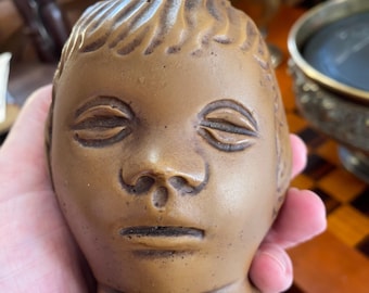 May include: A brown ceramic head sculpture with closed eyes, a small nose, and a defined mouth. The sculpture has detailed hair and is held in a hand. The head is a bust, with a flat base.