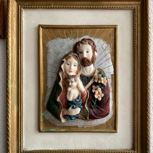 May include: A framed religious artwork depicting a holy family. The artwork features a three-dimensional sculpture of a man, woman, and child. The sculpture is painted in muted colors and has a textured surface. The frame is gold with a decorative border.