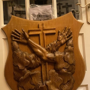 May include: A shield-shaped wooden wall plaque featuring a central carved cross and two raised arms reaching upward. The arms emerge from stylized carved clouds, with visible markings on the palms. The wood has a medium brown finish.