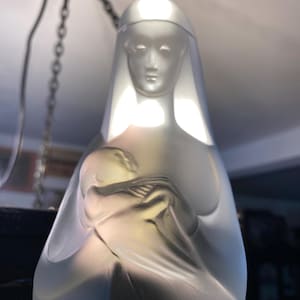 May include: A frosted glass sculpture of the Virgin Mary holding a baby Jesus. The sculpture is white and translucent, with a delicate, detailed design.