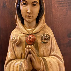 Puede incluir: Estatua beige de una mujer con cabello oscuro y una capa con capucha, sosteniendo una rosa roja. La estatua tiene dos rosas en la capa, sobre un fondo de madera oscura.
