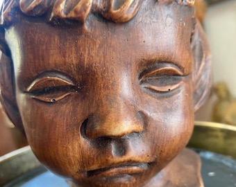 May include: A carved wooden bust of a child's face, featuring closed eyes and detailed hair. The sculpture is a warm brown color, with visible wood grain and texture. The bust is mounted on a small, geometric base.