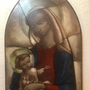 Puede incluir: Un panel de vidrieras que representa una escena religiosa. La obra de arte presenta a una mujer con una túnica azul y una prenda roja que sostiene a un niño con un halo. El panel tiene una parte superior redondeada y está compuesto por varias piezas de vidrio.