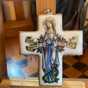 May include: A white cross-shaped pendant with an image of a woman in a blue and green robe, with a gold halo. The cross has the words "THE IMMACULATA" on it, with floral accents. The pendant is hanging on a wooden structure.