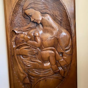 Może przedstawiać: Drewniana rzeźba ścienna przedstawiająca Matkę Boską trzymającą Dzieciątko Jezus. Rzeźba jest rzeźbiona w realistycznym stylu i charakteryzuje się skomplikowanymi detalami. Drewno ma bogaty brązowy kolor.