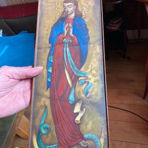 Può includere: Icona religiosa raffigurante una donna in abito rosso e mantello blu, con le mani giunte in preghiera. Un serpente verde con una mela rossa è in basso. Lo sfondo è dorato con stelle e l'opera è in una cornice rettangolare.