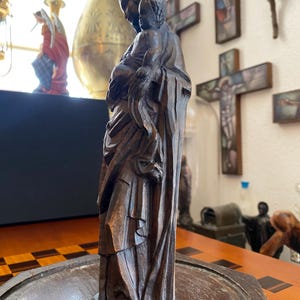 May include: A dark brown, carved wooden statue depicting a figure holding a smaller figure. The statue has detailed carvings of draped clothing and facial features. The figures are standing on a round base. The statue is likely religious in nature.