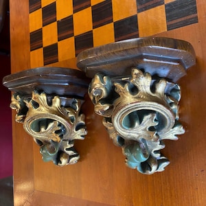 Puede incluir: Dos estanterías de pared antiguas de madera con detalles dorados y turquesas. Las estanterías tienen un acabado marrón oscuro y están decoradas con intrincados diseños de hojas y volutas. Las estanterías se muestran sobre una superficie de madera.