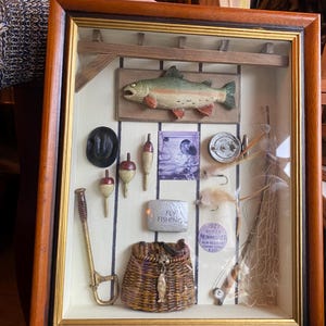 May include: A framed shadow box featuring fishing-themed items. Includes a wooden fish, bobbers, a fishing basket, and a net. The frame is brown with a gold interior border. The text "FLY FISHING" is visible on a metal plate.