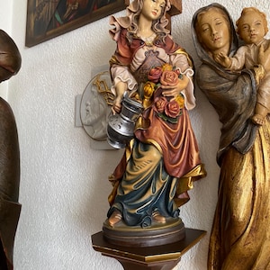 Może przedstawiać: Ręcznie malowana drewniana figurka kobiety w czerwono-niebieskiej szacie, trzymającej w jednej dłoni srebrzysty dzban, a w drugiej bukiet róż. Postać stoi na okrągłej podstawie umieszczonej na małej drewnianej półce ściennej.
