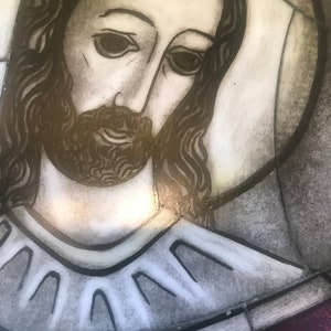 Puede incluir: Una representación de Jesucristo en vitral con cabello largo y oscuro y barba. Lleva una túnica blanca con un cuello decorativo. El fondo es gris y rojo apagado.