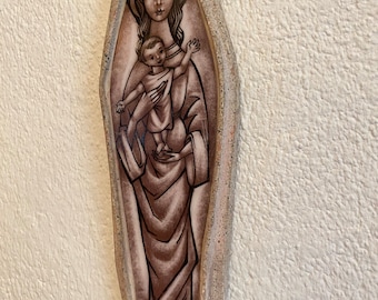 May include: A ceramic wall plaque depicting the Virgin Mary holding a baby Jesus. The plaque is in a brown and white colour scheme and has a textured surface.
