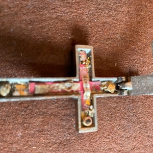 May include: A silver-toned metal crucifix pendant with a hinged design that opens to reveal a hollow interior. The inside features a red fabric lining, small metallic fragments, and a central vertical strip with text printed in black ink.