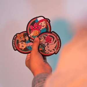 Könnte beinhalten: Drei runde Aufnäher mit Figuren und Kreaturen aus einer beliebten Science-Fiction-Serie. Jeder Aufnäher hat einen schwarzen Rand und ein farbenfrohes Design mit Text. Auf einem steht "Eleven vs Go", auf einem anderen "Save the Upside Down".