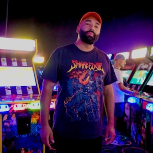 May include: A black short-sleeved t-shirt featuring a graphic print with the text "SNAKE EYES" in a stylized yellow font above a detailed illustration of a dark armored figure surrounded by orange and red flames, worn with a plain orange cap.