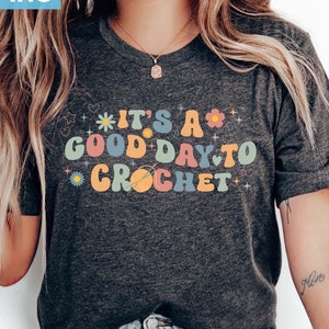 May include: A dark grey t-shirt with the text "It's a good day to crochet" in colorful letters with flowers and stars. The shirt is being worn by a person with long blonde hair and blue jeans.